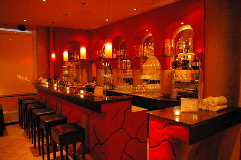 Barbereich mit roter Theke, schwarzen Hockern und beleuchteten Flaschenregalen.